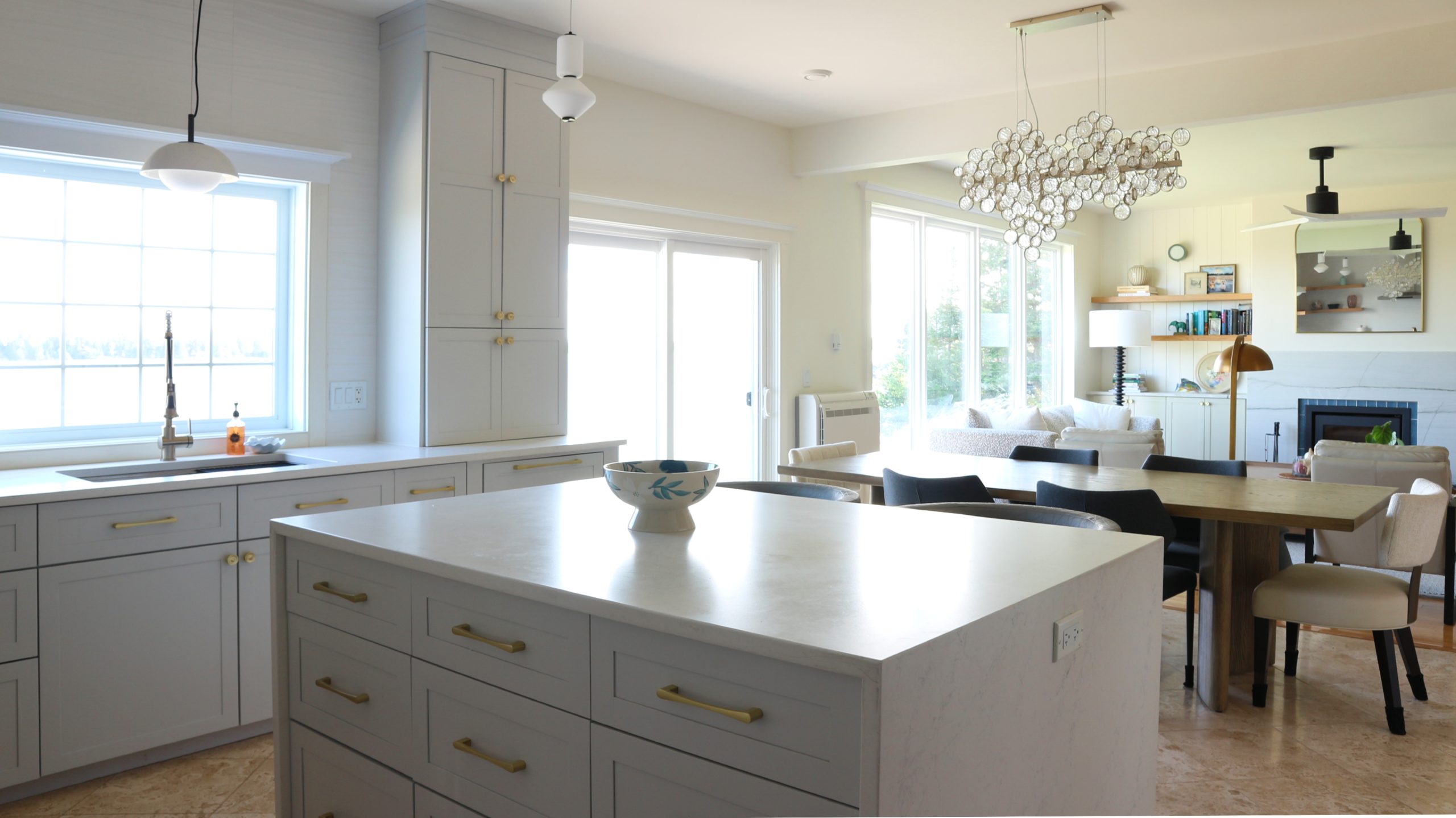 pale grey kitchen cabinets with gold pulls and glass and ceramic light fixtures and dining and living areas beyond